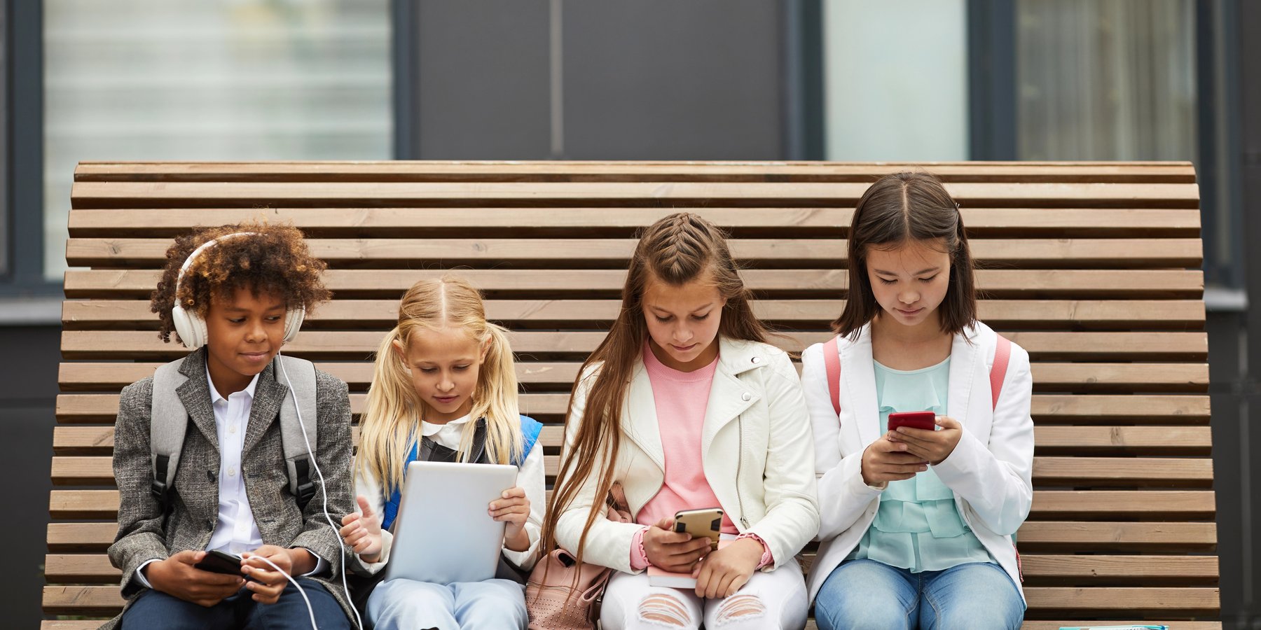 Four children sitting together outside, each absorbed in a different device